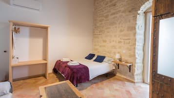 A bedroom with a stone wall, a bed with a purple blanket, a wooden nightstand with a lamp, and a wooden shelf with clothes.