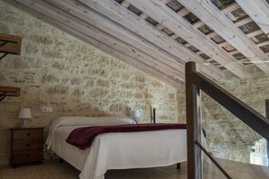 Ein Schlafzimmer mit einer Steinsäule, einem Bett mit weißer Bettdecke, einem Nachttisch aus Holz mit einer Lampe und einer Holztreppe.