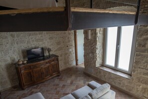 Une pièce avec un mur en pierre, un meuble en bois sur lequel est posée une télévision, une fenêtre et un canapé.