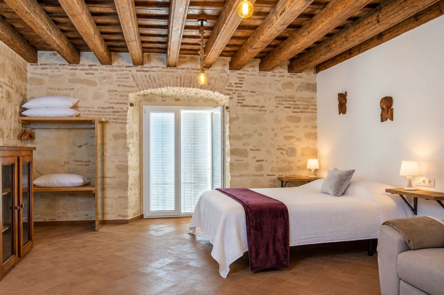 Un dormitorio con pared de piedra, vigas de madera, cama con almohadas, mesita de noche y una puerta con persianas.