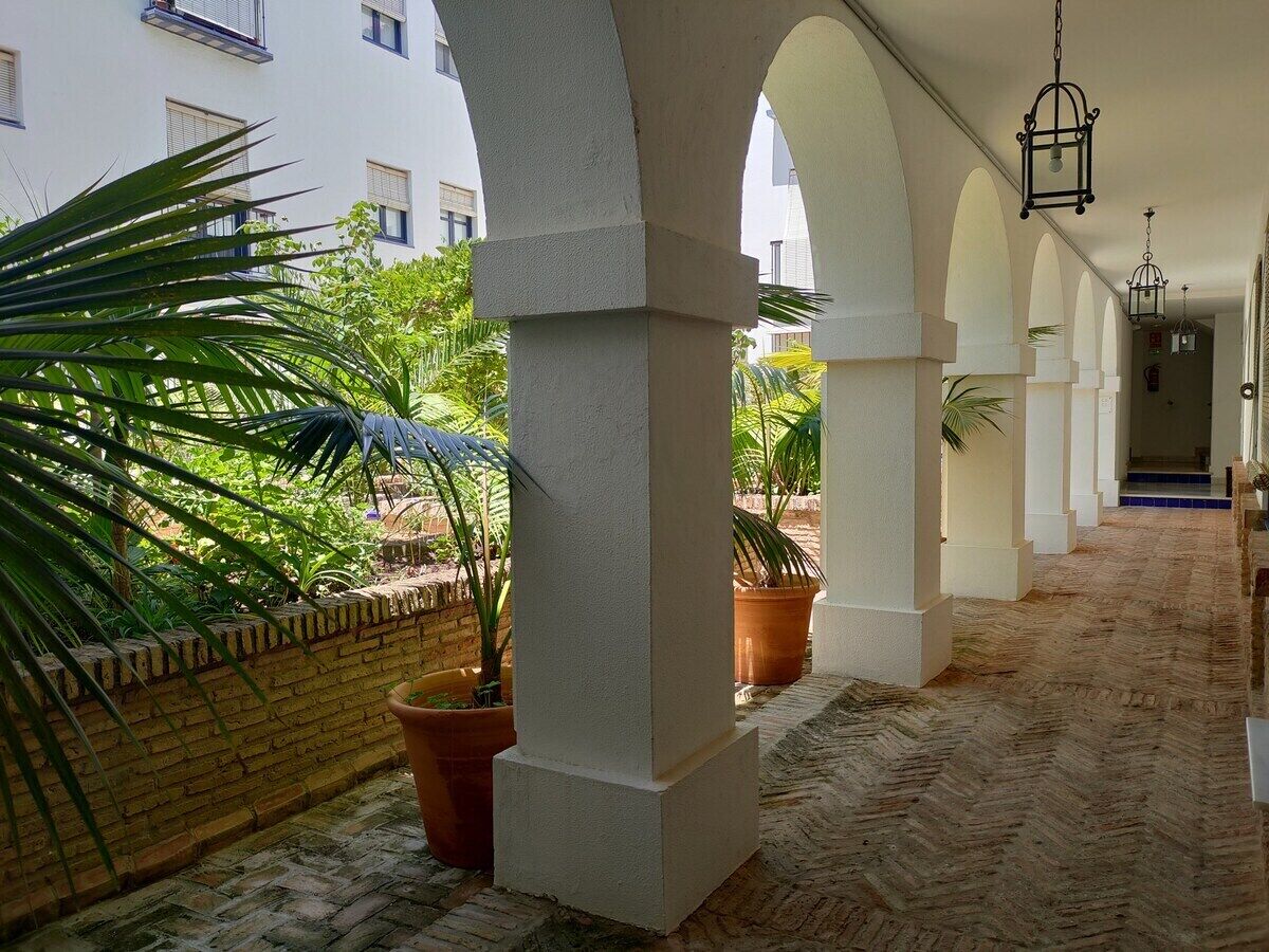 A covered outdoor corridor with arches, potted plants, and a brick wall.