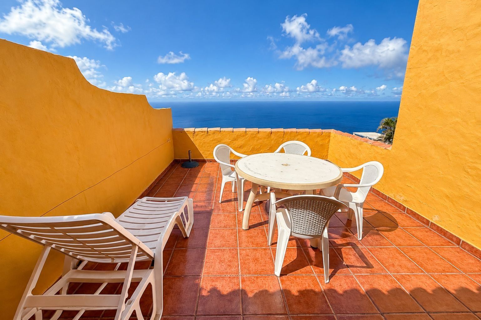 A terrace with a round table and white chairs, overlooking a coastal view.