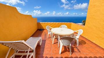 A terrace with a round table and white chairs, overlooking a coastal view.
