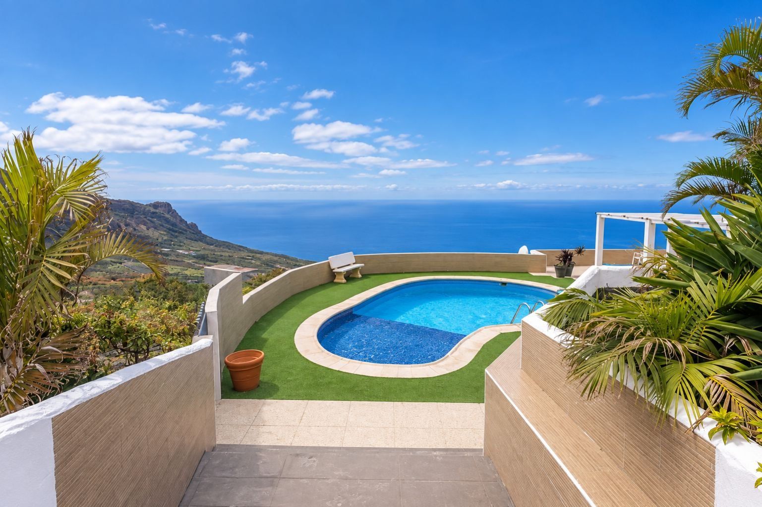 A curved swimming pool with a view of the ocean, surrounded by greenery and a white structure.