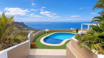A curved swimming pool with a view of the ocean, surrounded by greenery and a white structure.