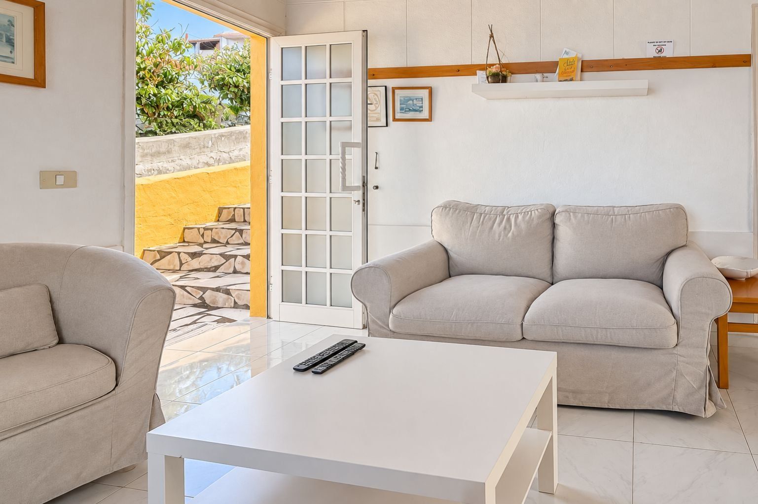 Una sala de estar con un sofá color beige, una mesa de centro blanca y una puerta que da a un área exterior.