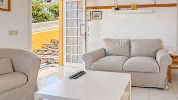 Una sala de estar con un sofá color beige, una mesa de centro blanca y una puerta que da a un área exterior.