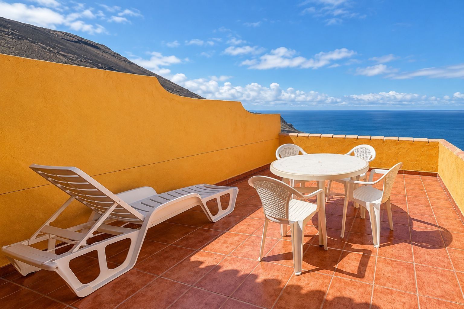 Una terraza con mesa y sillas, una tumbona y vista al mar.