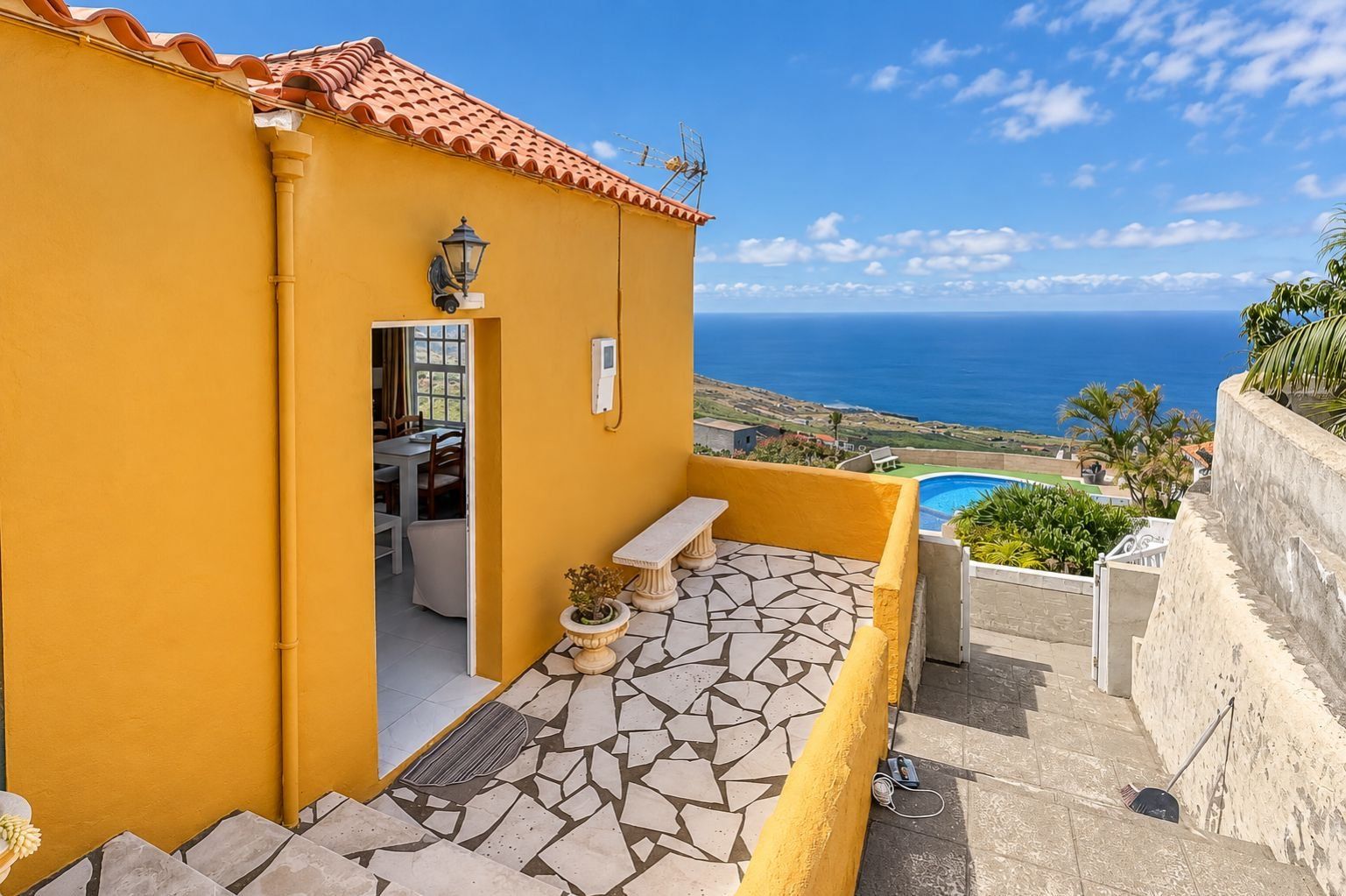 Un edificio amarillo con patio empedrado, piscina y vista al mar.