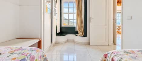 Quarto de hotel com duas camas, uma janela com cortinas, uma porta e vista para o exterior.