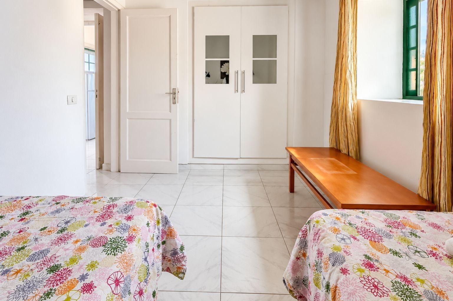 Una habitación con dos camas, un armario, un banco y una ventana con cortinas.