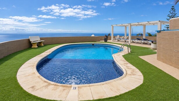 Ein Pool mit Meerblick, umgeben von Kunstgräsern und Liegestühlen.