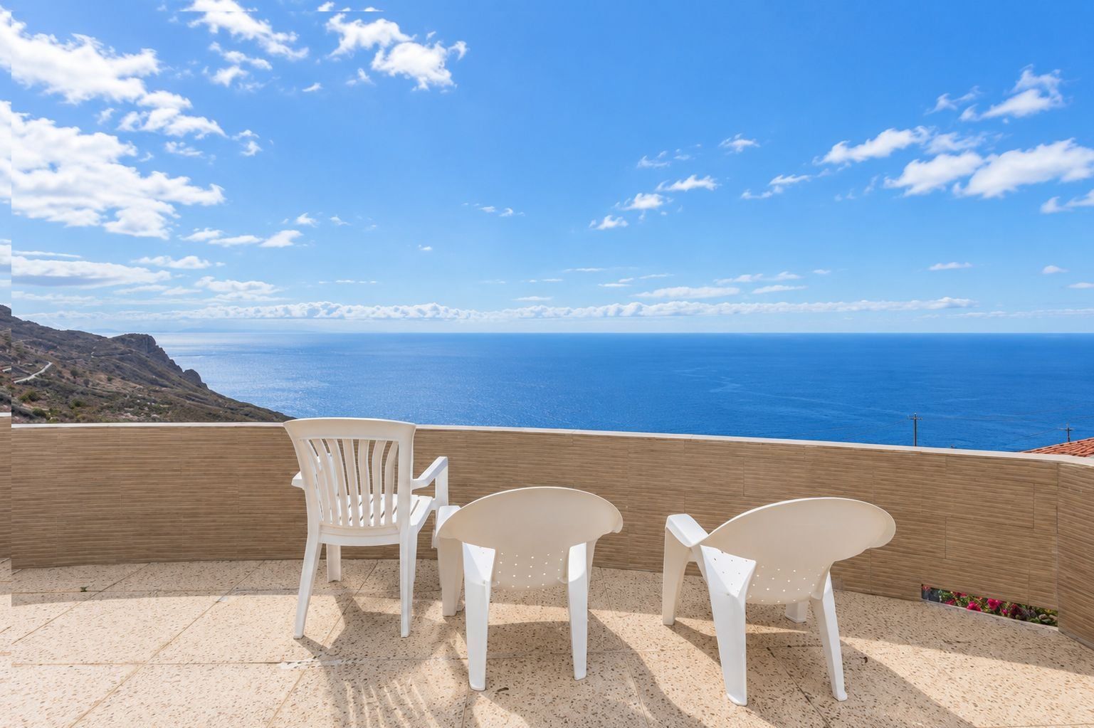 Una terraza con tres sillas blancas y una mesa, con vistas a un paisaje costero bajo un cielo despejado.