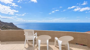 Una terraza con tres sillas blancas y una mesa, con vistas a un paisaje costero bajo un cielo despejado.