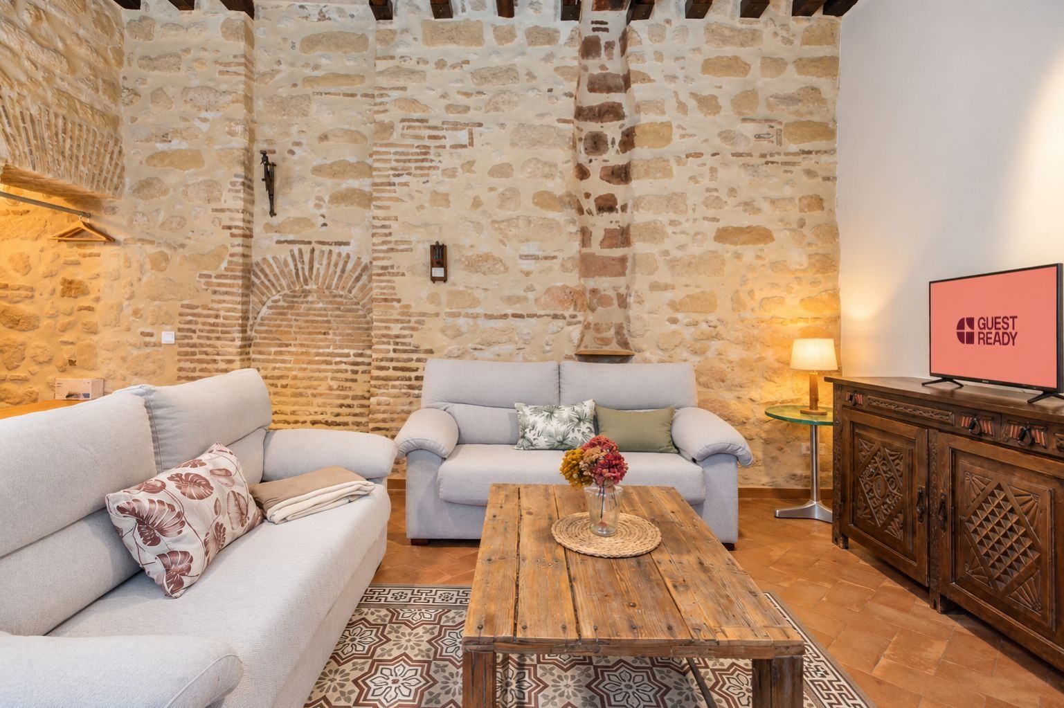 Une pièce chaleureuse aux murs de pierre, avec deux canapés, une table basse en bois et un téléviseur affichant le message « PRÊT POUR LES CLIENTS ».