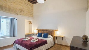 Un dormitorio con pared de piedra, una cama con cabecero de madera, dos mesitas de noche con lámparas y una ventana con cortinas.