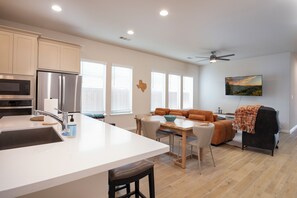 A modern kitchen with a central island, dining area with a table and chairs, a sofa, and a ceiling fan. - Luxe Home, King Bed, Yoga/Office Room, Private Backyard, Community Pool! (Little Elm)