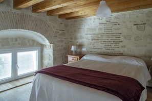 A bedroom with a stone wall, a bed with a white and maroon bedspread, a nightstand with a lamp, and a window with blinds. - GuestReady - La Cruz 2C (El Puerto de Santa María)