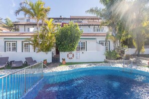 Una piscina con borde de azulejos, rodeada de sillas de descanso y palmeras, con un edificio al fondo.