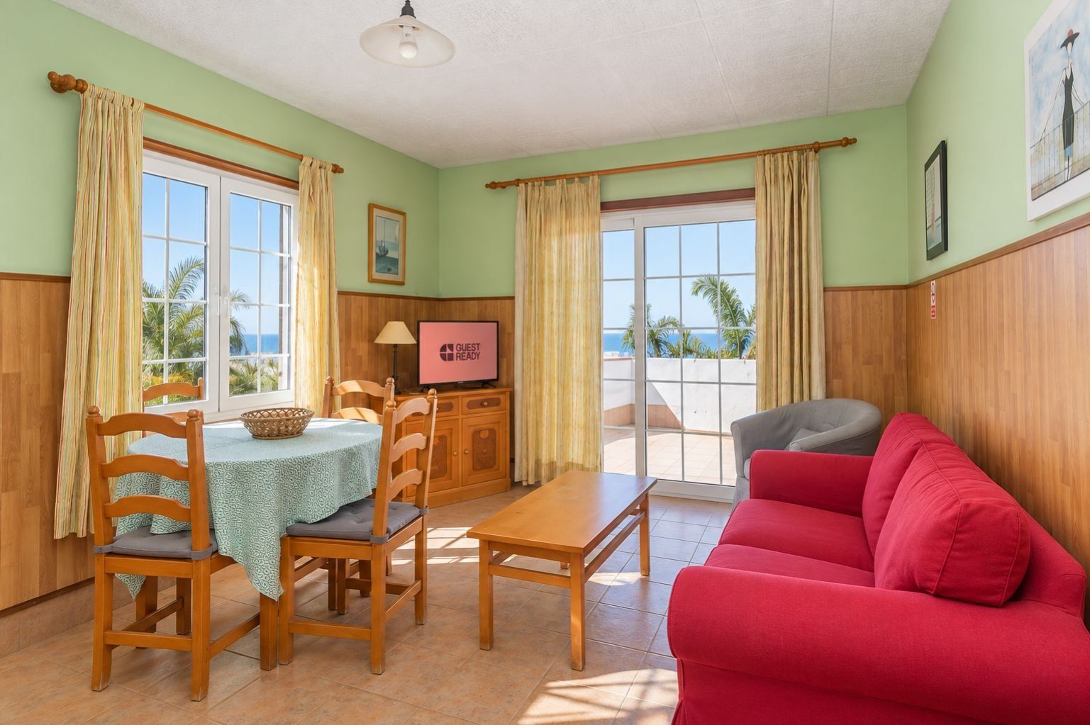 Ein Zimmer mit einem Esstisch, Stühlen, einem roten Sofa, einem Fernseher und Blick aufs Meer.