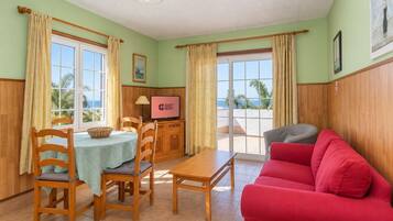 Ein Zimmer mit einem Esstisch, Stühlen, einem roten Sofa, einem Fernseher und Blick aufs Meer.