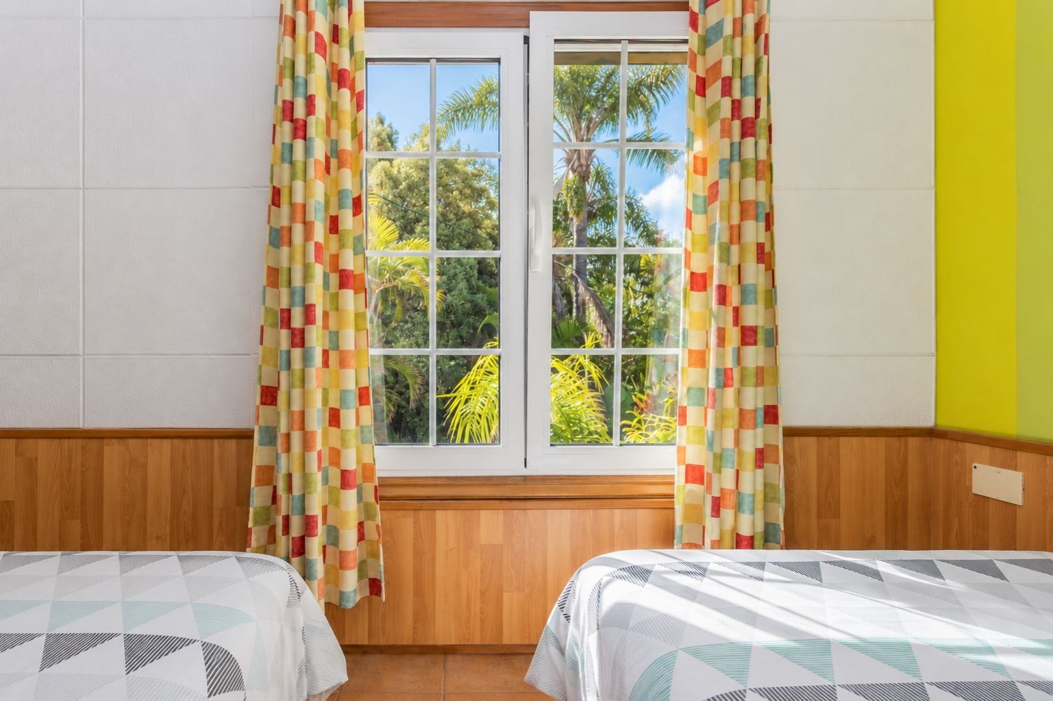 Una habitación con dos camas, una ventana con cortinas, paredes de madera y vistas a zonas verdes.