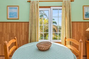 Ein Zimmer mit Holzboden, einem runden Tisch mit gemustertem Tischläufer, zwei Holzstühlen, einem gewebten Korb und einem Fenster mit Vorhängen.