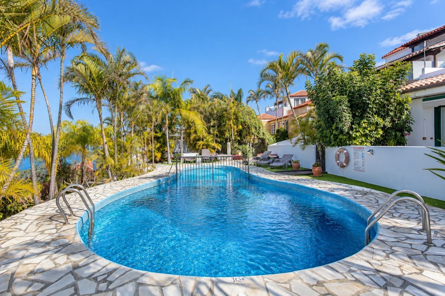 Una piscina con borde de azulejos, rodeada de palmeras y un edificio blanco con techo rojo.