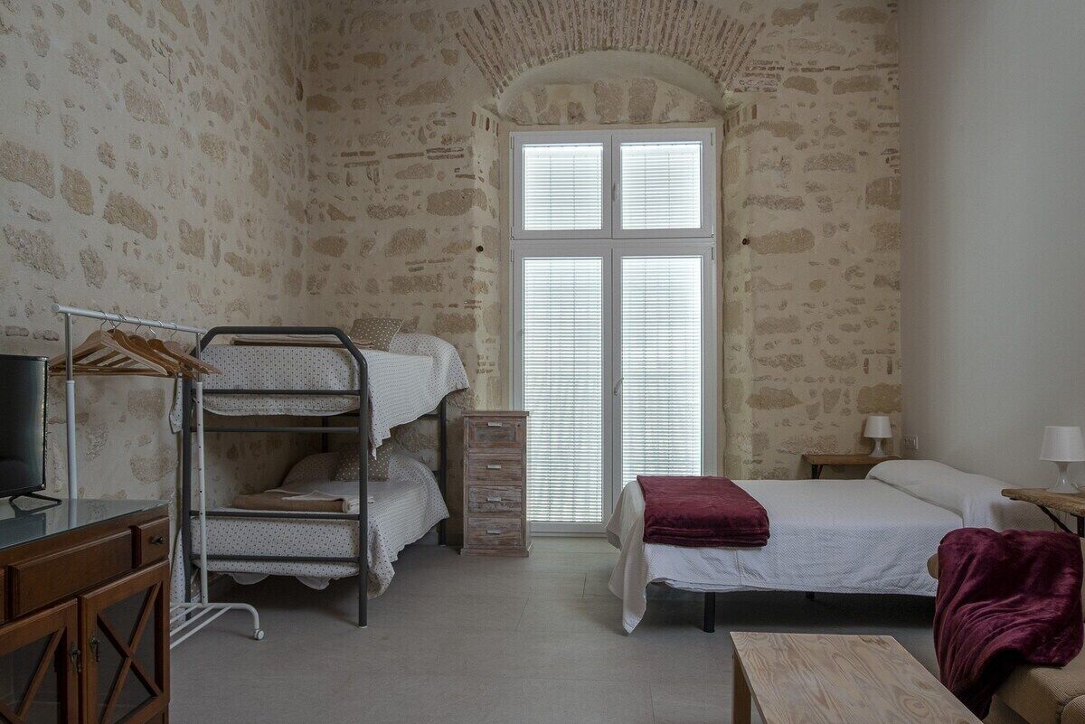 A room with bunk beds, a desk with a television, a wooden dresser, and a window with blinds.