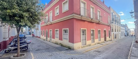 A red building with a balcony, a cobblestone street, and parked scooters.