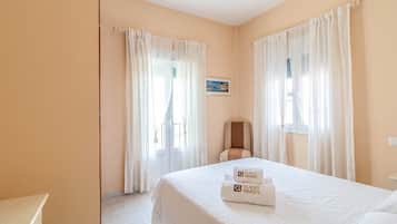 Habitación de hotel con cama, dos ventanas con cortinas, una silla y un cuadro en la pared.