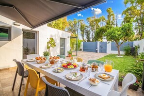 Un espace de restauration en plein air avec une table dressée pour le petit-déjeuner, entourée de chaises, et donnant sur un jardin avec des arbres et une clôture.