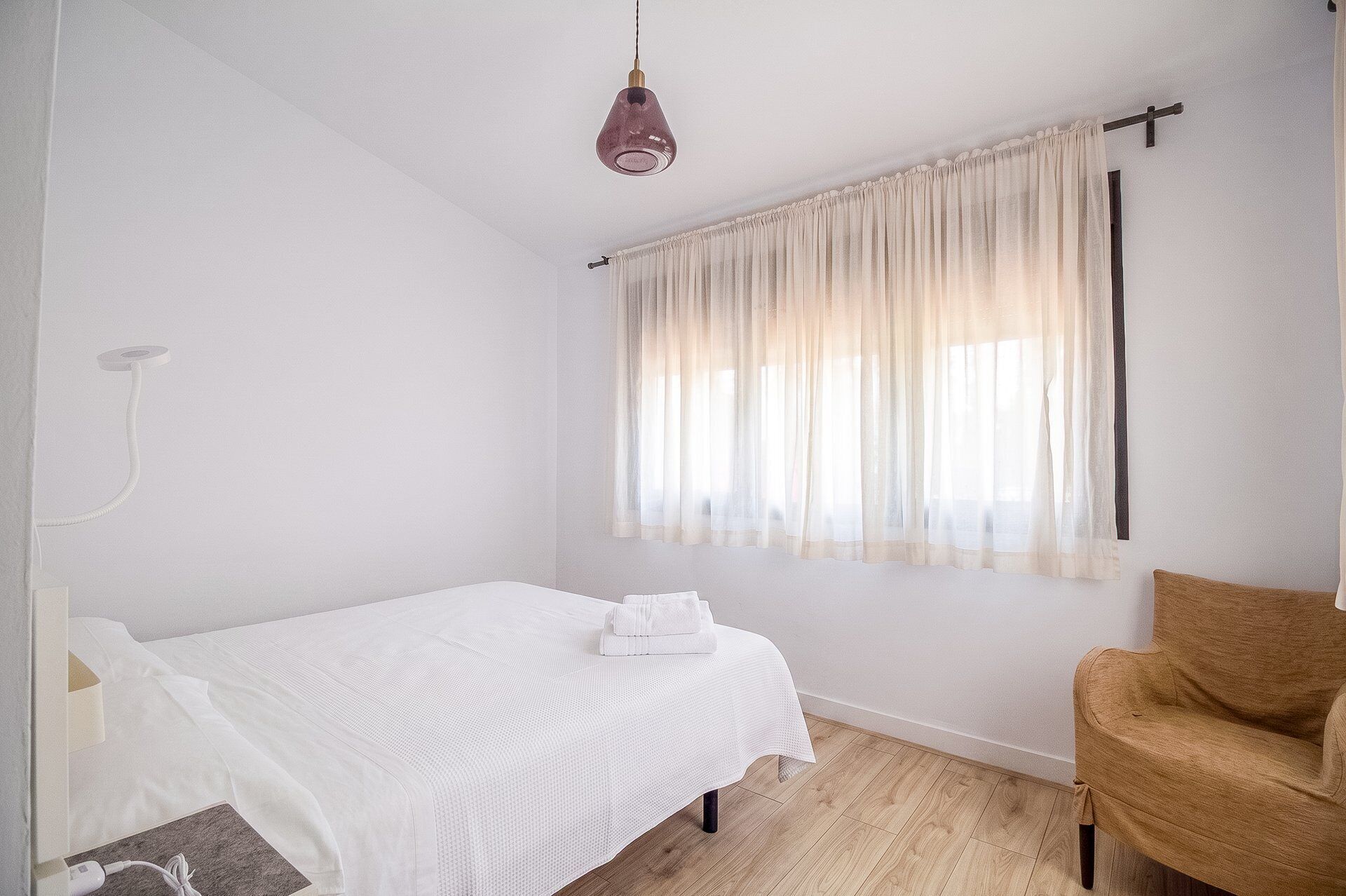 Ein schlicht eingerichtetes Schlafzimmer mit einem Einzelbett, einem Nachttisch, einem Sessel und einem Fenster mit leichtem Vorhang.