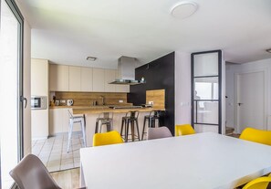 A modern kitchen with a central island, bar stools, and a dining table with yellow chairs. - GuestReady - Valdelagrana Gus (El Puerto de Santa María)