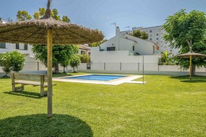 A pool area with a thatched umbrella, a wooden bench, and a white building in the background. - GuestReady - Valdelagrana Gus (El Puerto de Santa María)