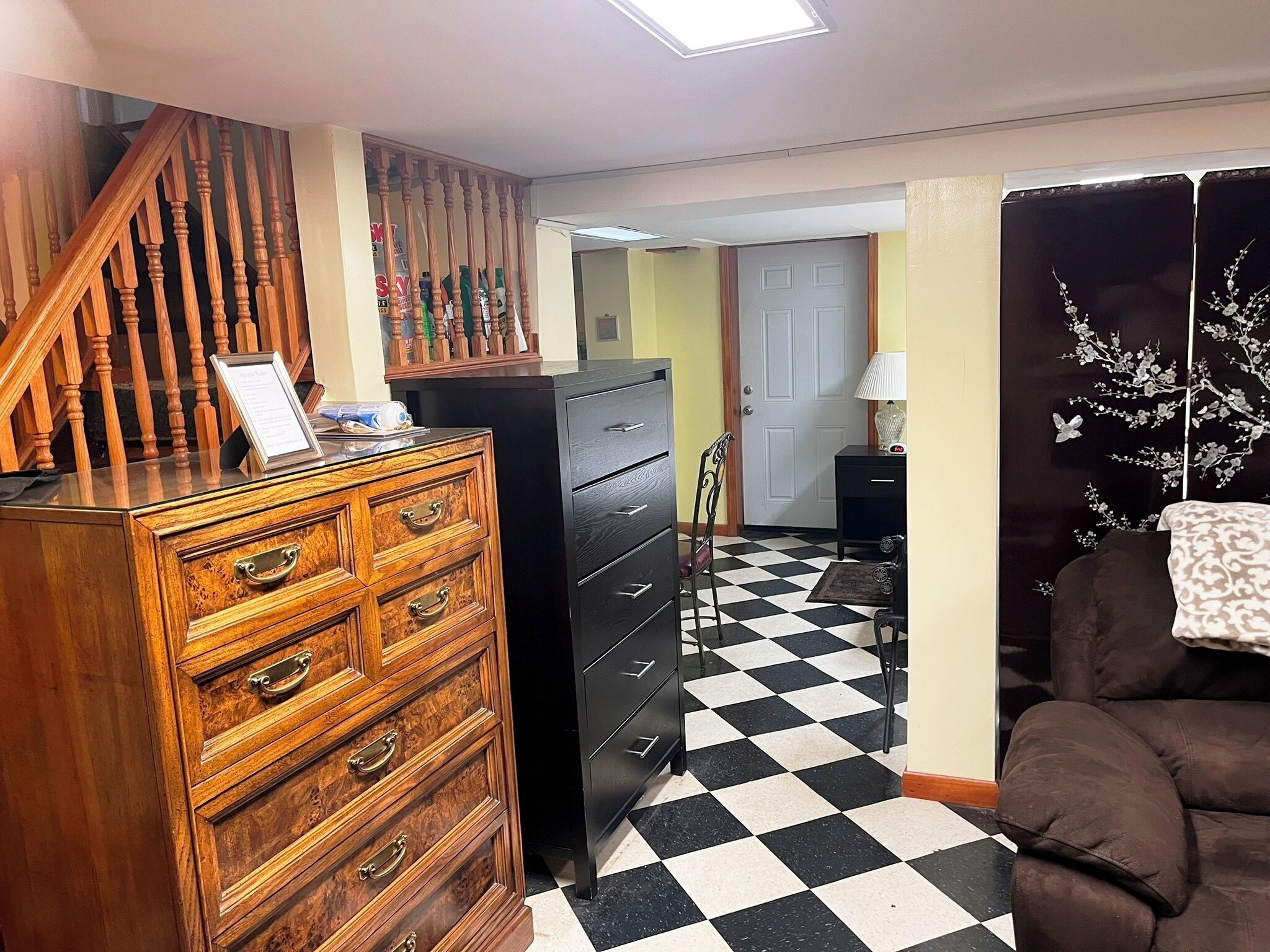 A room with a staircase, a wooden dresser, a black filing cabinet, a checkered floor, and a brown sofa.