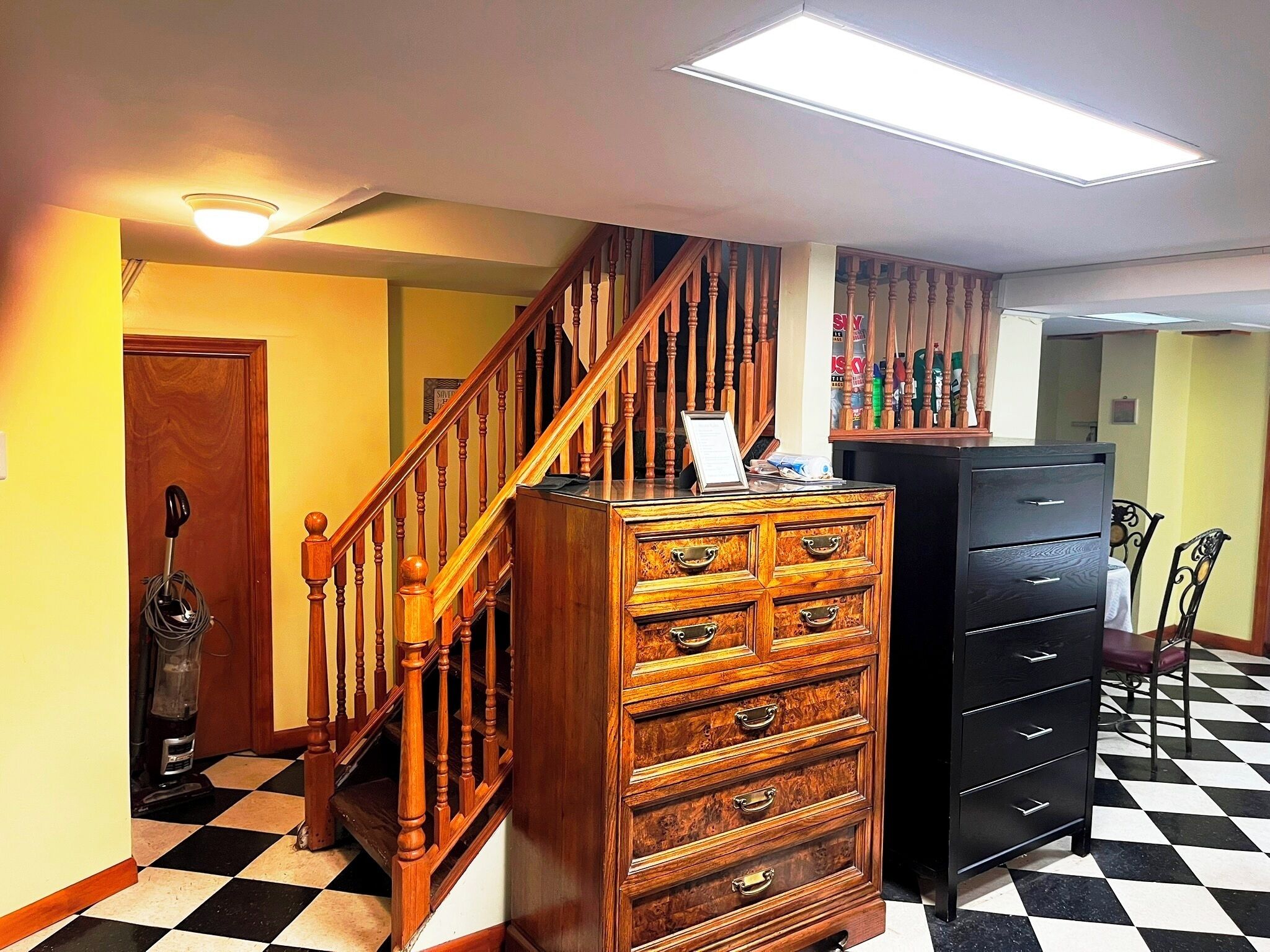 A staircase with wooden balusters, a wooden chest of drawers, a black filing cabinet, and a checkered floor.