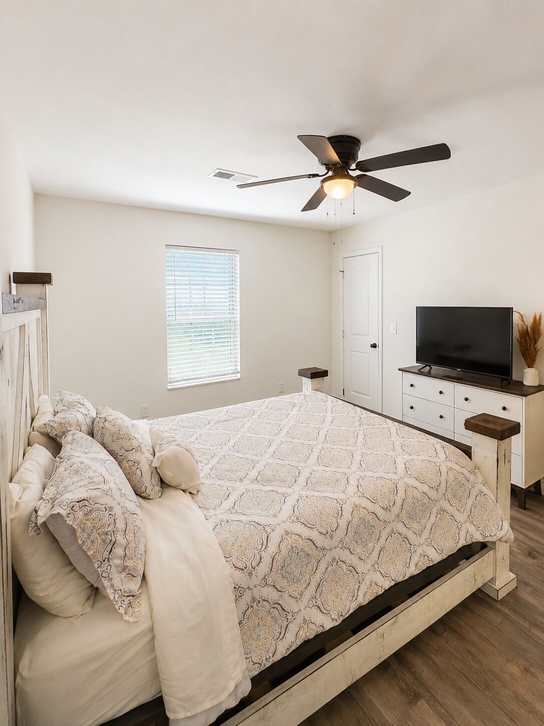 A bedroom with a bed, a ceiling fan, a television, and a window with blinds.