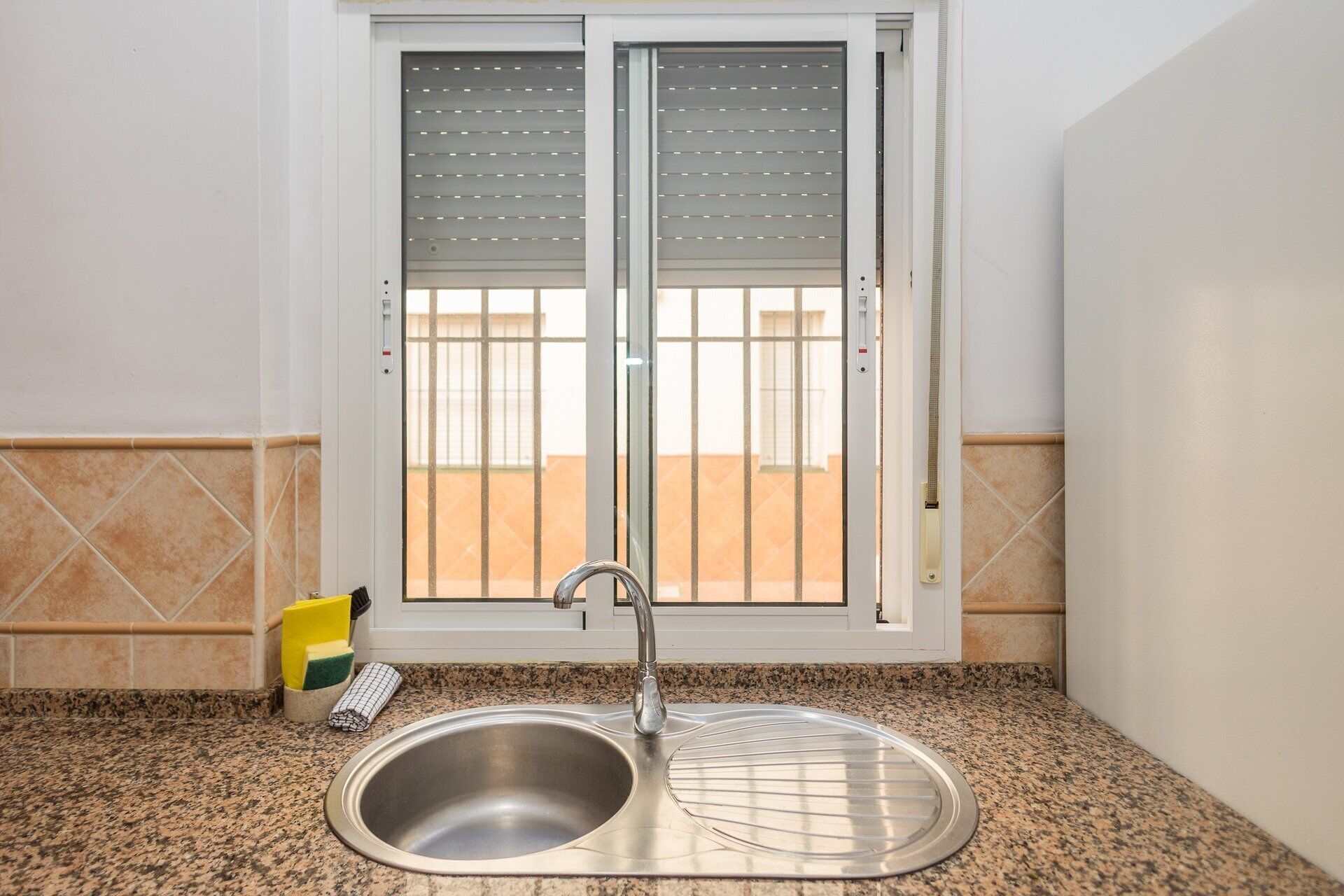 Una cocina con un lavamanos de acero inoxidable, una ventana con persianas y un salpicadero de azulejos.
