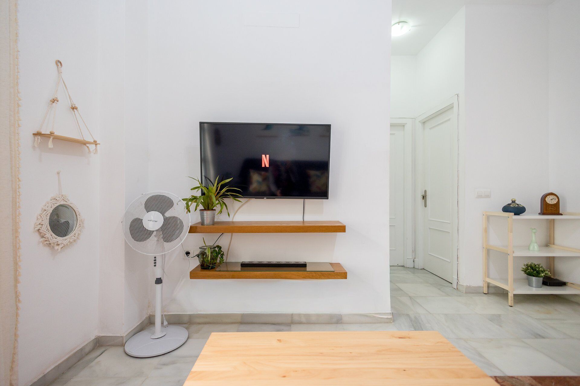 A living room with a flat-screen TV mounted on the wall, a standing fan, a wooden coffee table, and a potted plant.