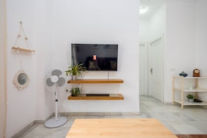Una sala de estar con una televisión de pantalla plana montada en la pared, un ventilador de pie, una mesa de centro de madera y una planta en maceta.