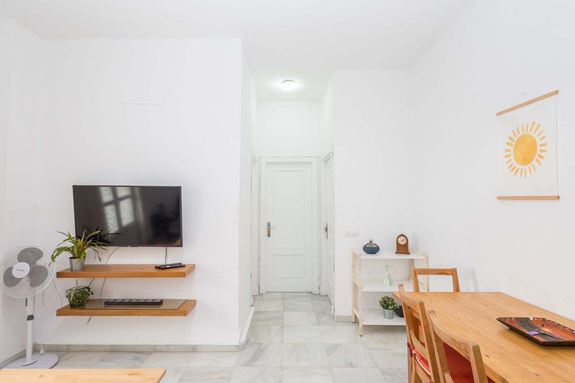 Una sala moderna con televisor de pantalla plana, estanterías de madera, un ventilador, una mesa de comedor y una puerta que da a otra habitación.