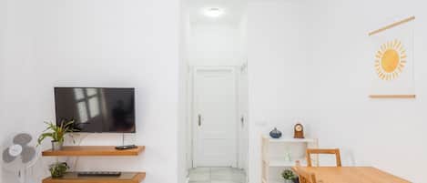 A modern living room with a flat-screen TV, wooden shelves, a fan, a dining table, and a door leading to another room.