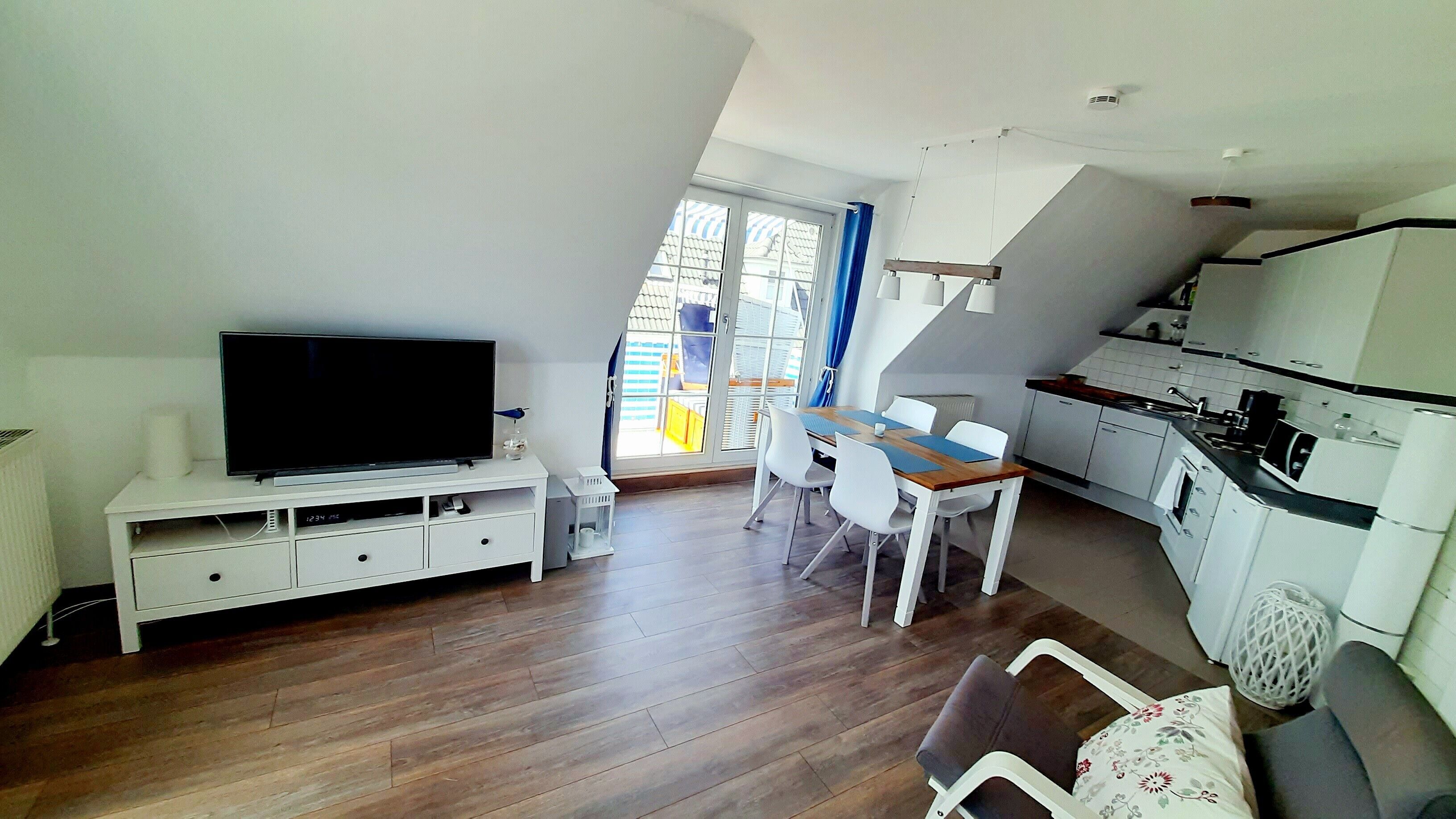 Un espace de vie compact comprenant un téléviseur posé sur un meuble blanc, une table à manger avec des chaises, un coin cuisine et une vue par la fenêtre.