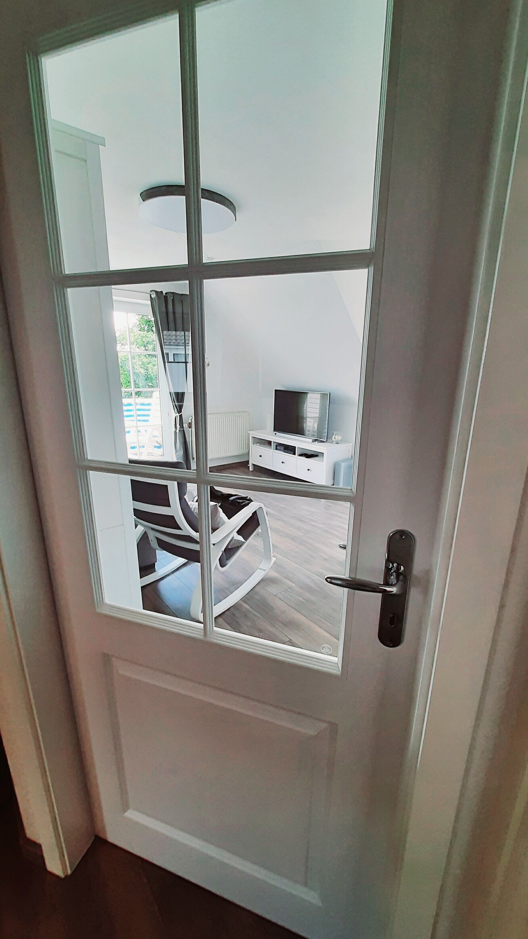 Eine Tür mit Glasfenster, ein heller Innenraum, ein Fernseher auf einem weißen Schrank und ein Bett mit weißem Kopfteil.