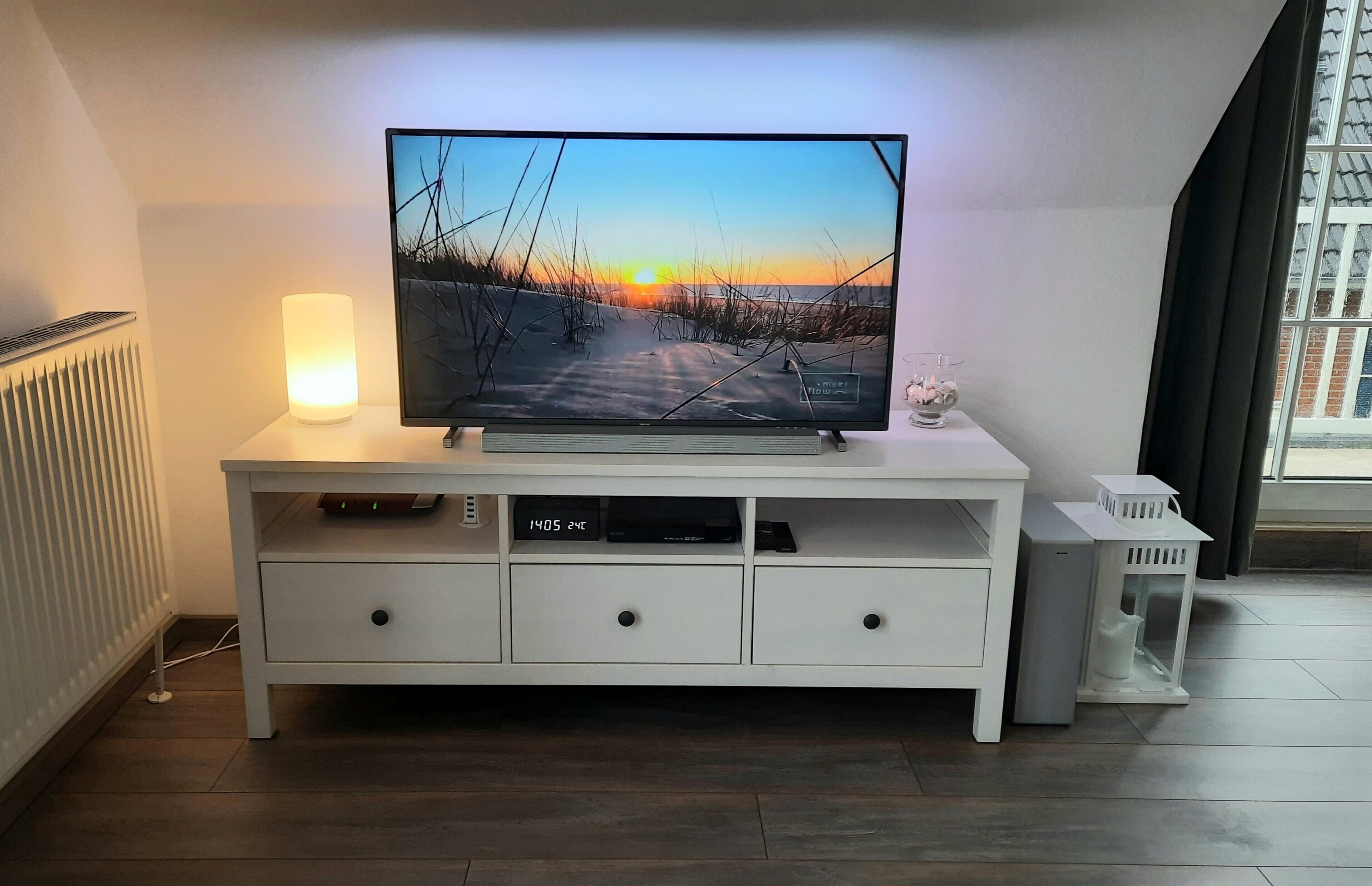 Uma sala de estar moderna com uma televisão de tela plana sobre um móvel branco, um abajur de chão e uma janela com vista para um edifício.