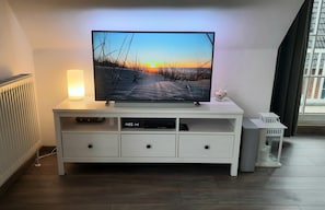 Ein modernes Wohnzimmer mit einem Flachbildfernseher auf einer weißen Unterhaltungsanlage, einer Stehlampe und einem Fenster mit Blick auf ein Gebäude.