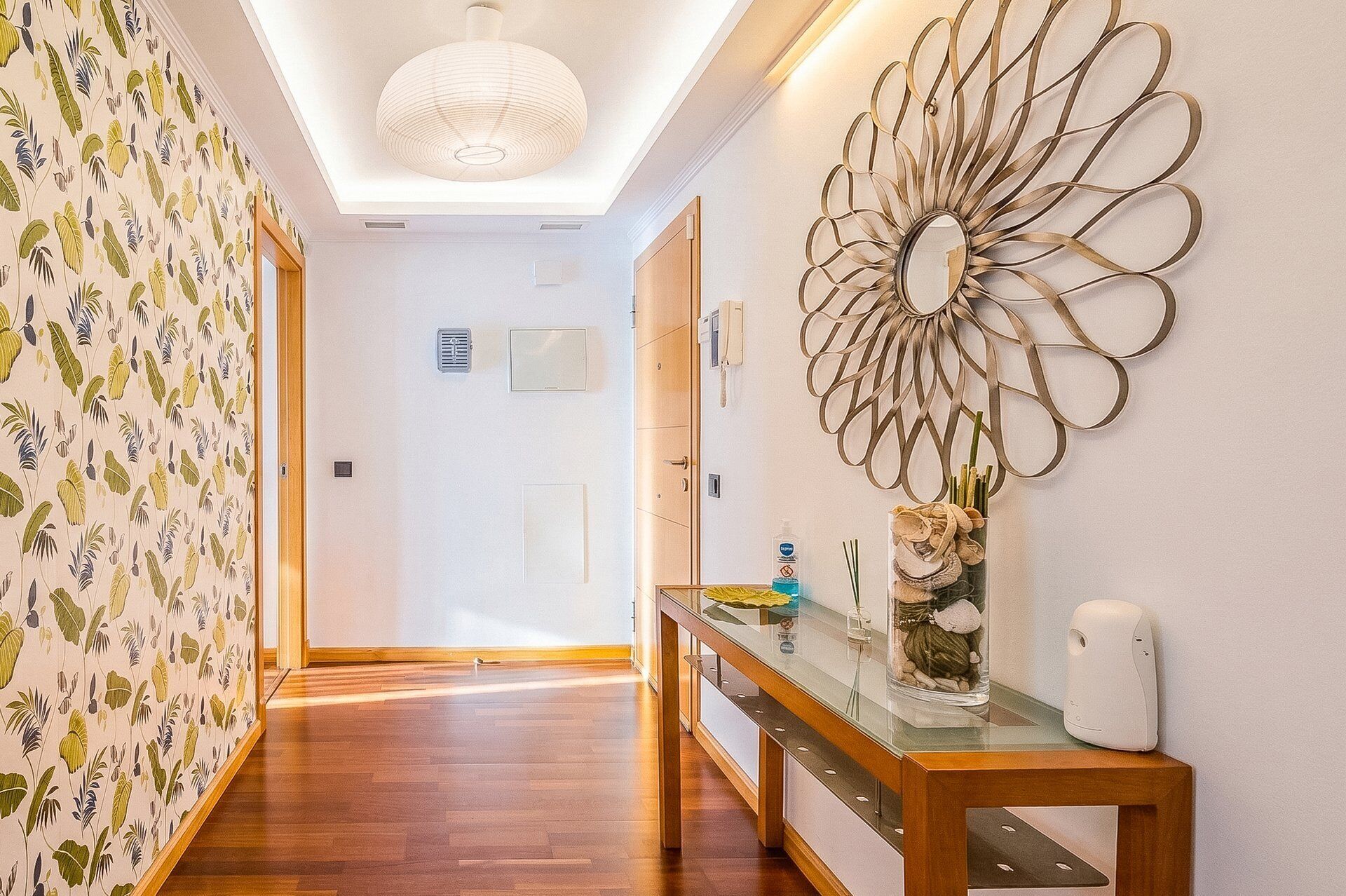 A hallway with a wooden floor, a decorative wall piece, and a glass-top console table with a vase and a bottle.
