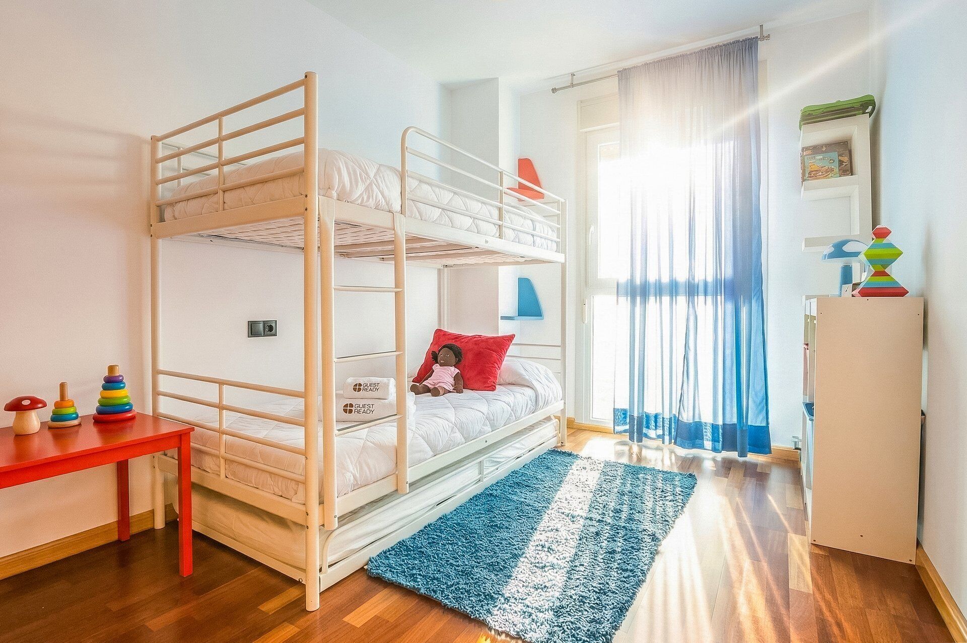 Une chambre avec un lit superposé, une table rouge, un tapis bleu et une fenêtre avec des rideaux.
