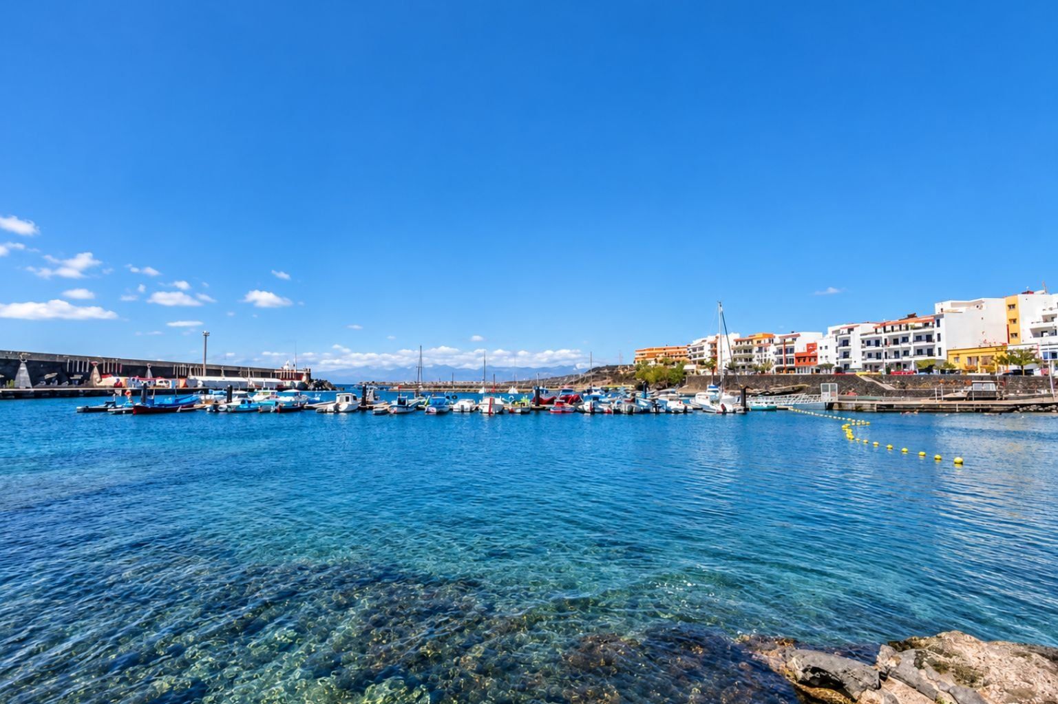 A marina with boats docked, clear water, and buildings in the background.
